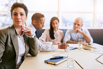 Single out of a mature businesswoman seated in front of her three colleagues sitting behind her still busy working as she smiles confidently at the camera.