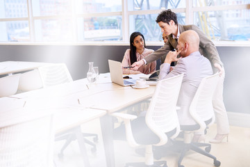 Fototapeta premium Mature female supervisor pointing at a laptop screen with a pen during a meeting between three of her employees in a bright modern conference room with a large window behind them.