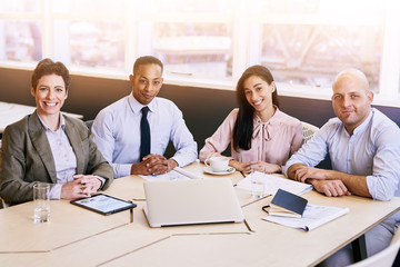 4 business professionals all looking at the camera during a business meeting in a modern conference room with a large window behind them with bright natural light coming in.