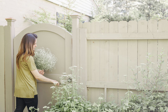 Woman Entering A Garden Through A Gate, Carrying A Bunch Of White Flowers.