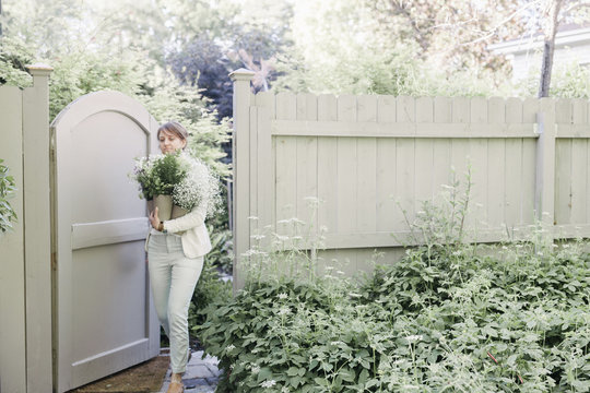 Woman Entering A Garden Through A Gate, Carrying A Bunch Of White Flowers.
