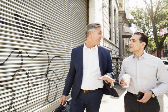 Two men walking along a street past a steel shutter on a building, painted with graffiti. 