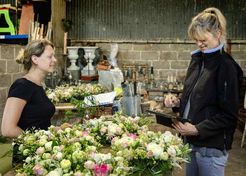 Two women, one using a digital tablet. Florists at a workbench covered in table decorations, pink and white flower arrangements. 