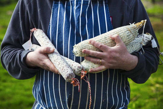Close up of a butcher wearing a striped blue apron, standing outdoors, holding a selection of salamis.