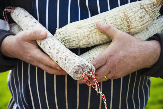 Close up of a butcher wearing a striped blue apron, standing outdoors, holding a selection of salamis.