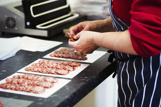 Close up of a butcher wearing a striped blue apron, packaging slices of salami.