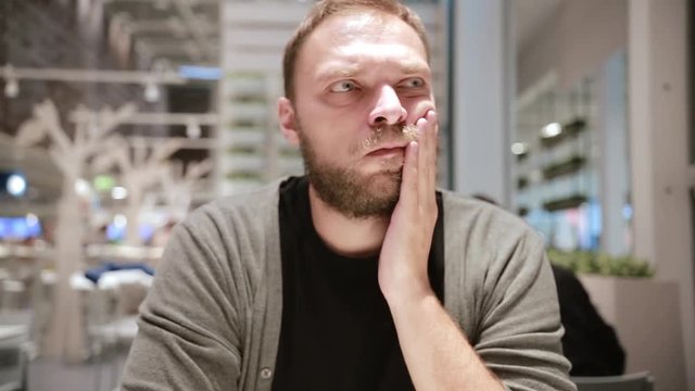 Serious handsome man with a beard and grey touched his face sitting in the cafe, toothache.