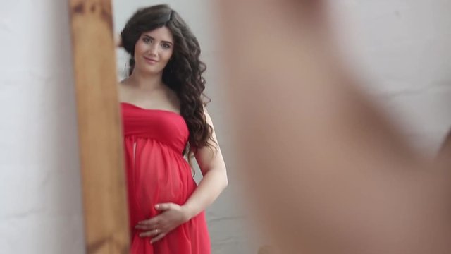 Pregnant brunette woman in a red dress supports stomach looking at the mirror