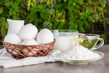 Пшеничная мука, куриные яйца, подсолнечное масло и чашка с молоком на деревянном столе
