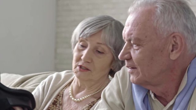 Elderly couple sitting on sofa holding virtual reality glasses and discussing them, then senior woman putting them on