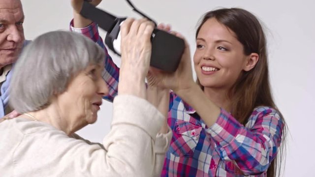 Young woman in virtual reality glasses sitting with her grandparents, then putting VR headset on excited senior woman and laughing at her reaction