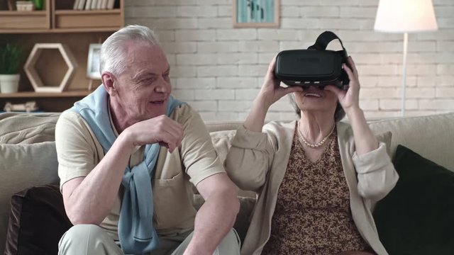 Widely smiling elderly woman looking into virtual reality glasses and sitting on sofa beside senior man