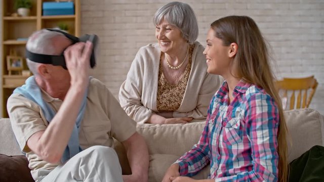 Elderly man in virtual reality headset looking around room in amazement as laughing young woman and senior lady waving hands before him to check if he can see them