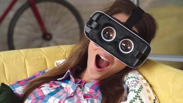 Young woman lying on sofa wearing virtual reality goggles without front panel, then making silly face and looking at camera in slow motion