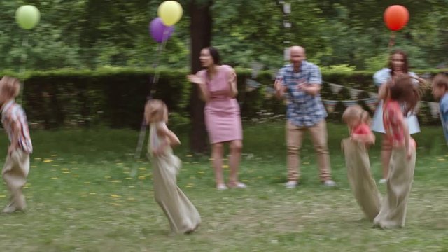 Little boys and girls jumping in sacks through the green lawn in the park and falling on their way while their parents standing and clapping hands