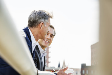 Two people, men talking together outdoors leaning on a railing, taking a coffee break. 