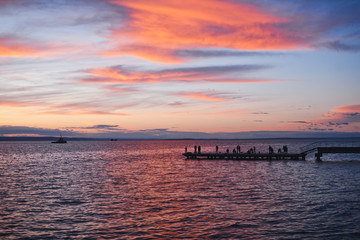 Fototapeta na wymiar Bright sunset on seaside with pier, fishermen and ships silhouette. Cloudy sky at sunset painted with pink and orange colors