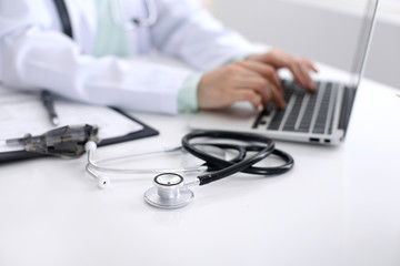 Close-up of stethoscope is lying on the table near female doctor typing on laptop computer
