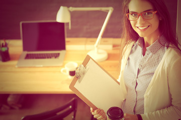 Young woman standing near desk with laptop holding folder and cup of coffee