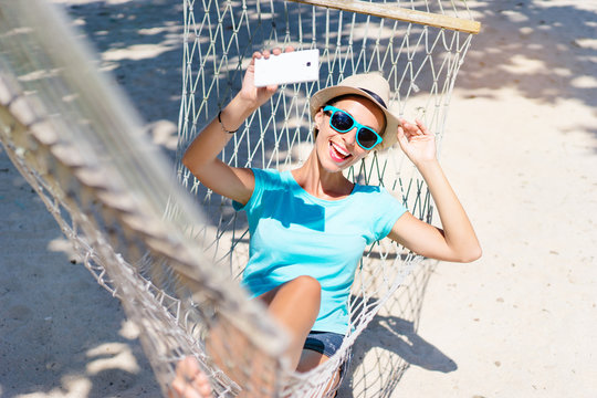 Vacation selfie. Enjoying the summer. Young pretty woman in hat and sunglasses using smartphone laying in hammock on the beach.
