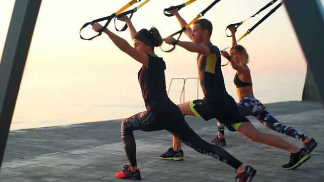 Beautiful sportive people training with trx at seaside at sunrise. Slow motion.