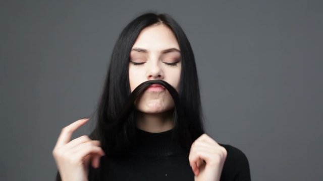 Young brunette woman is playing with long hair and making mustache of it, gray background