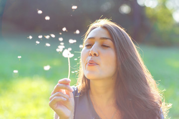 Beautiful young woman blowing a dandelion in park.