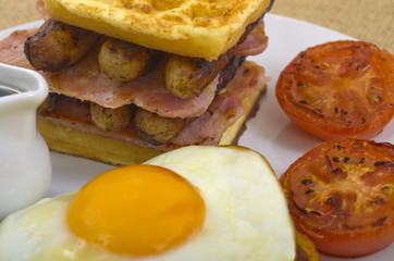 Waffle Stack Breakfast
Breakfast of waffles, bacon, egg, sausage in a stack,  a side of tomato, and sunny side up egg sat on the hash browns with a jug of maple syrup