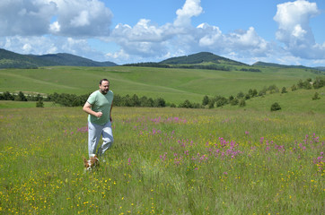Fototapeta na wymiar Man and his dog running in a field with wildflowers on a cloudy day in springtime and with hills in background