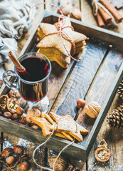 Glass of mulled wine in wooden tray with Christmas gingerbread cookies, nuts, cinnamon, anise and pine cones over rustic wooden background, selective focus, copy space, vertical composition