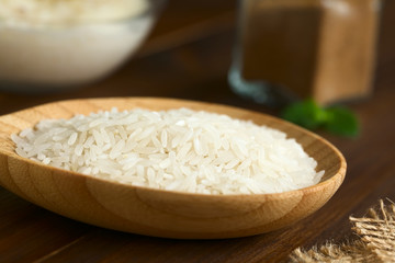 Raw white rice grains on small wooden plate, cinnamon powder and a bowl of rice pudding in the back, photographed with natural light (Selective Focus, Focus one third into the rice grains)