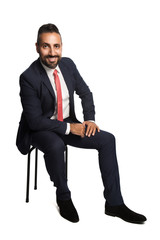 An attractive and smiling businessman wearing a blue suit and red tie, sitting down on a chair with hand on his knee. White background.