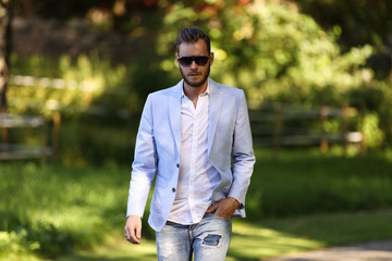 An attractive man wearing a light blue blazer, white shirt and sunglasses, walking along a path...
