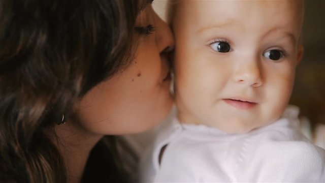 Young Happy Mother Holding her Newborn Child and kissing. Family at home. Beautiful smiling Mom and Happy Baby Together. Tenderness concept.