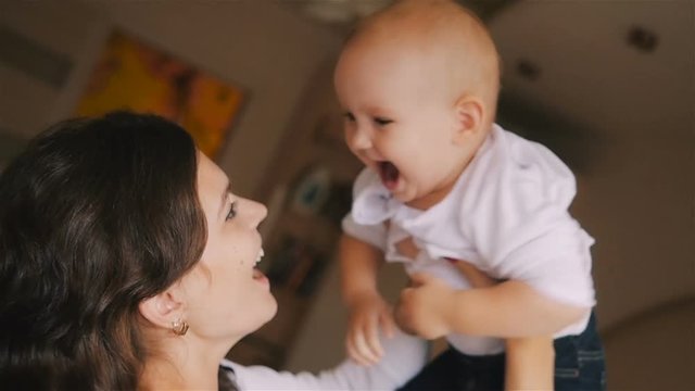 Young Happy Mother Holding her Newborn Child and throws up. Family at home. Beautiful smiling Mom and Happy Baby Together in bed.