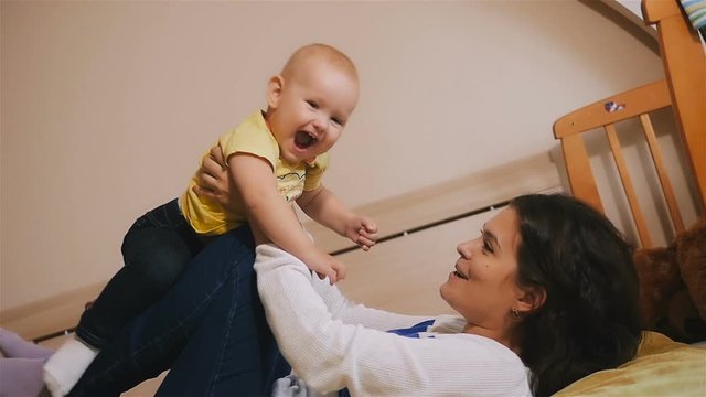 Young Happy Mother Holding her Newborn Child and throws up. Family at home. Beautiful smiling Mom and Happy Baby Together in bed.