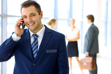 Smiling businessman  in office with colleagues in the background and using mobile