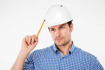 Thoughtful young man builder in building helmet with pencil thinking