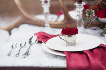 Wedding table setting in marsala colors with plates, cutlery, red floral compositions, candles, velvet napkins, deer horns on table covered with snow