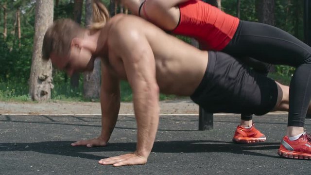 Shirtless muscular man doing push-ups on the park playground with woman on his back