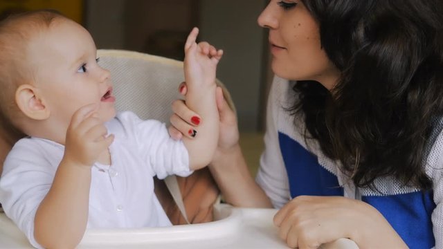 Young Happy Mother Playing with her Newborn Child while seating in baby seat. Family at home. Beautiful smiling Mom and Happy Baby Together.