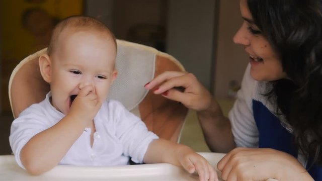 Young Happy Mother Playing with her Newborn Child while seating in baby seat. Family at home. Beautiful smiling Mom and Happy Baby Together.