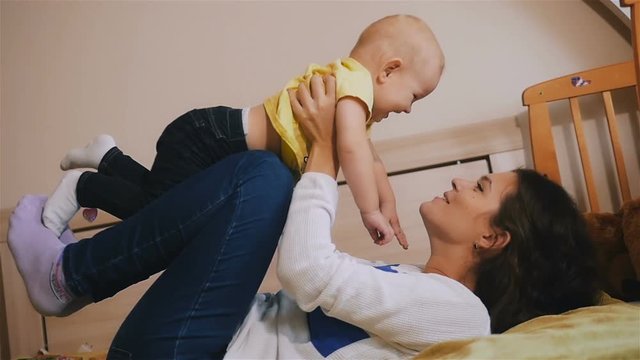 Young Happy Mother Holding her Newborn Child and throws up. Family at home. Beautiful smiling Mom and Happy Baby Together in bed.