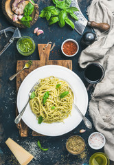 Italian pasta dinner. Spaghetti with pesto sauce and fresh basil, Parmesan cheese and spices served with glass of wine on rustic wooden board over dark grunge plywood background, top view