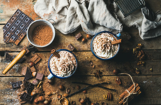 Hot chocolate with whipped cream, nuts and cinnamon in enamel mugs with ingredients around on rustic wooden background, top view, horizontal composition