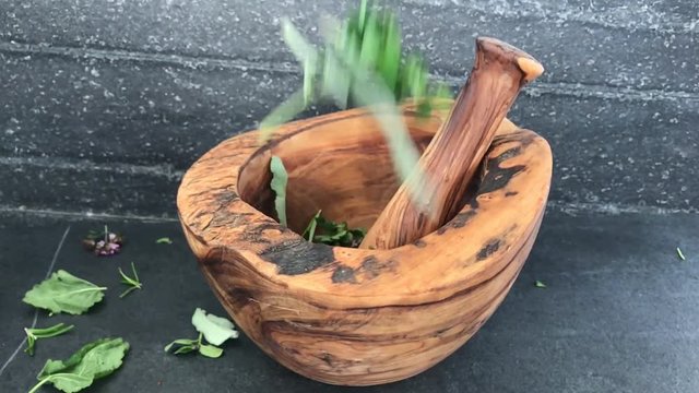 Fresh herbs flying into olive wood mortar & pestle, black stone background in the background. Slow motion.