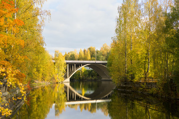 Bridge over still water with colorful trees on the sides. Image taken during beautiful autumn day in Finland.