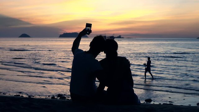 Silhouette of couple taking selfie photo sitting on beach during sunset
