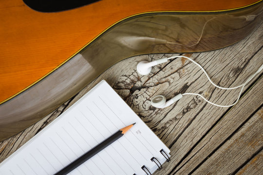 Black pencil with notebook, white headphones and guitar on wooden background