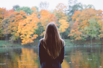 Outdoor autumn portrait of girl standing backwards with straight hair. Back view of woman explores calm lake and forest nature at fall. Image toned with old film style filters